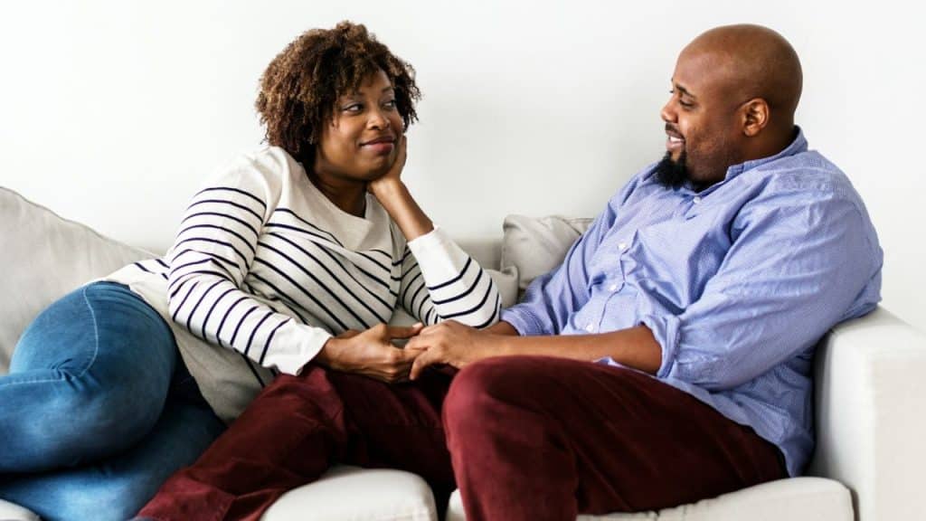 A Black couple is seated on a white couch, looking at each other. The woman is resting her chin on her hand.