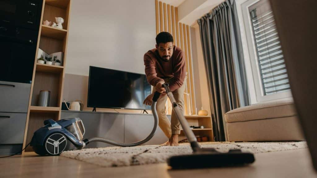 Man with dark hair vacuuming a living room carpet with a canister vacuum cleaner.