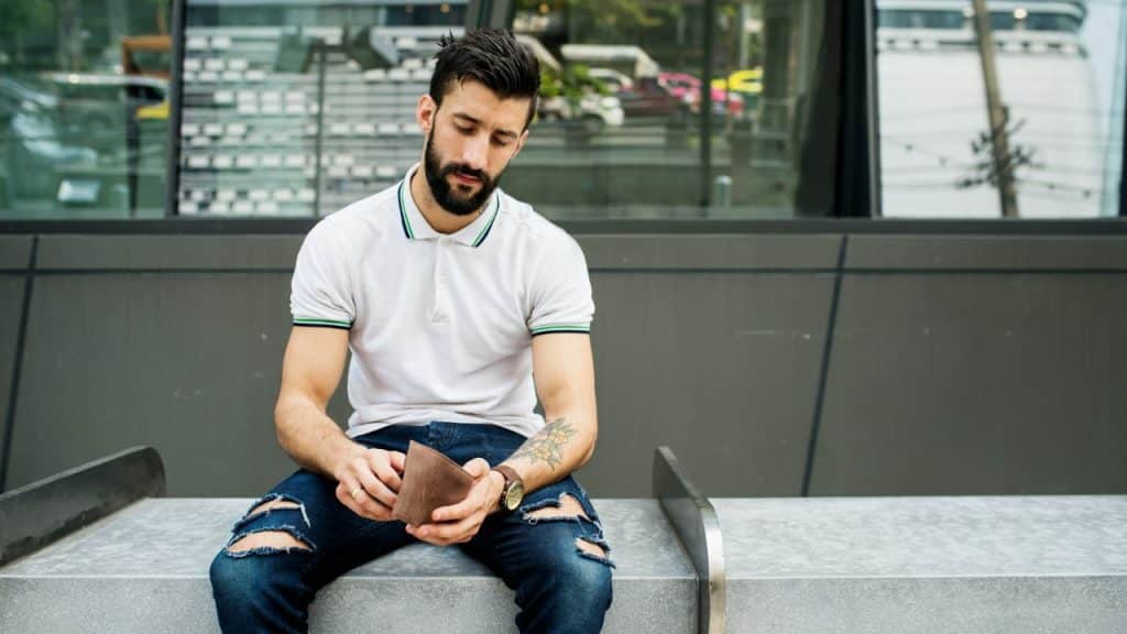 A bearded man in a white polo shirt and ripped jeans sits outdoors looking down at an empty brown wallet.
