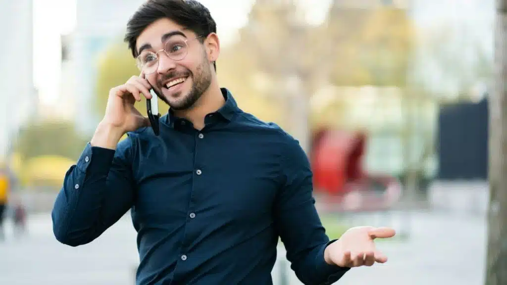 A smiling man with round glasses talks on a phone outdoors, gesturing with his hand.