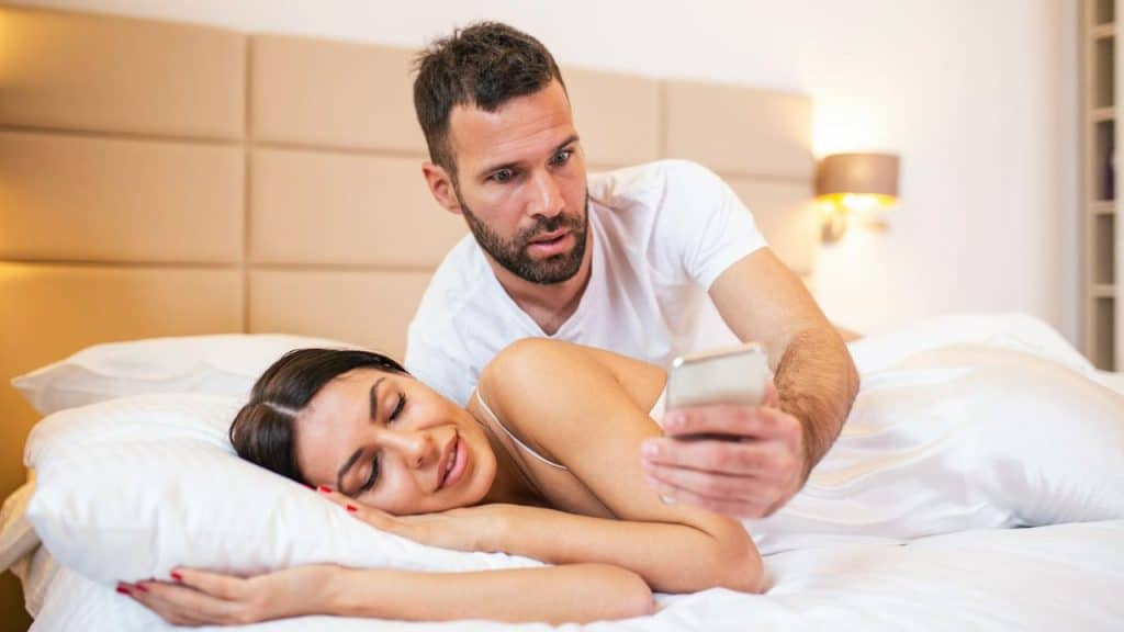 A serious man leans over a woman sleeping in bed and holds her smartphone to look at it.