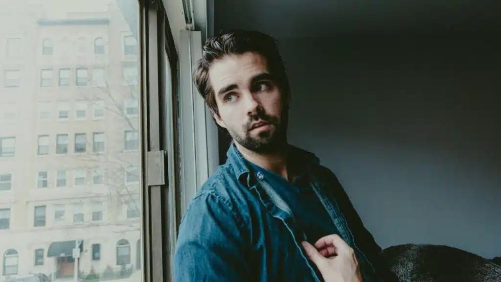 Man in a denim shirt looking out a window at a brick building.