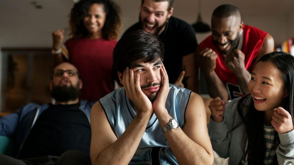 A sad man in a light blue jersey is sitting with five happy, diverse adults cheering around him.