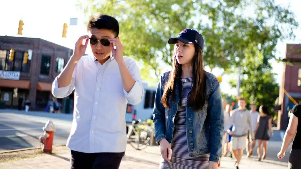 A man is adjusting his sunglasses while walking next to a  woman in a baseball cap on an urban sidewalk.