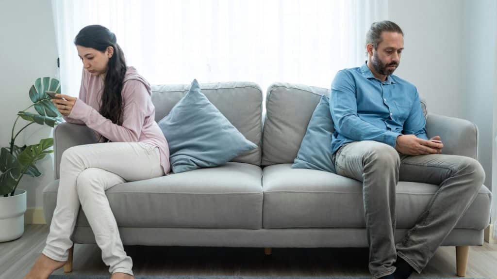 A man and a woman sit far apart on a gray couch, separated by a blue throw pillow. The woman looks at her phone.