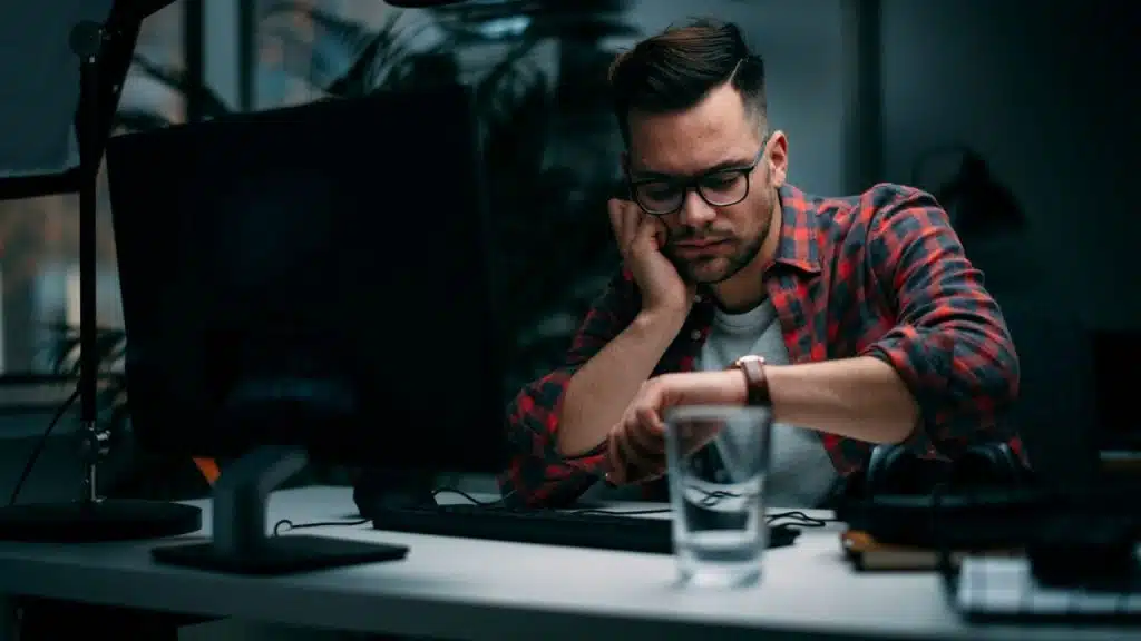 A man in a plaid shirt and glasses looks at his wristwatch while sitting at a computer.