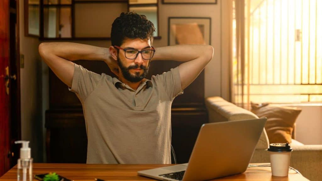 Man with glasses and a beard sits at a desk with a laptop, stretching his arms behind his head.
