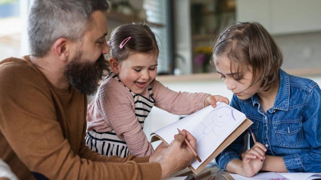 Smiling man with a beard draws with two young girls at a table.