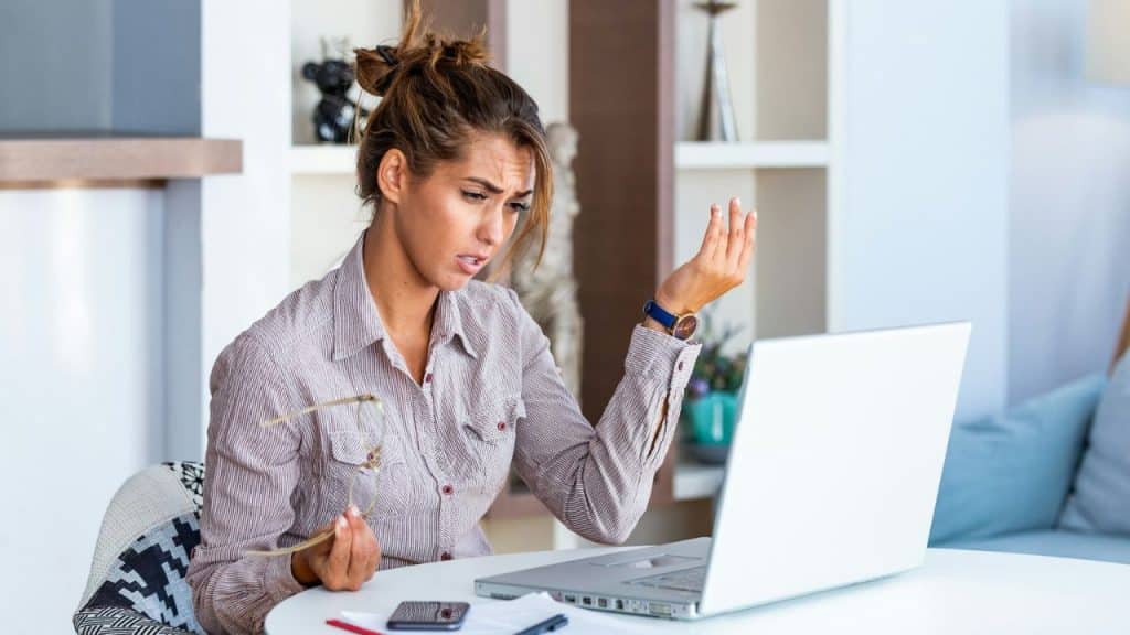 Frowning woman at a laptop holds glasses in one hand, gesturing with the other.