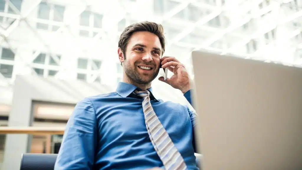 A man in a blue shirt smiling while talking on his phone and using a laptop.