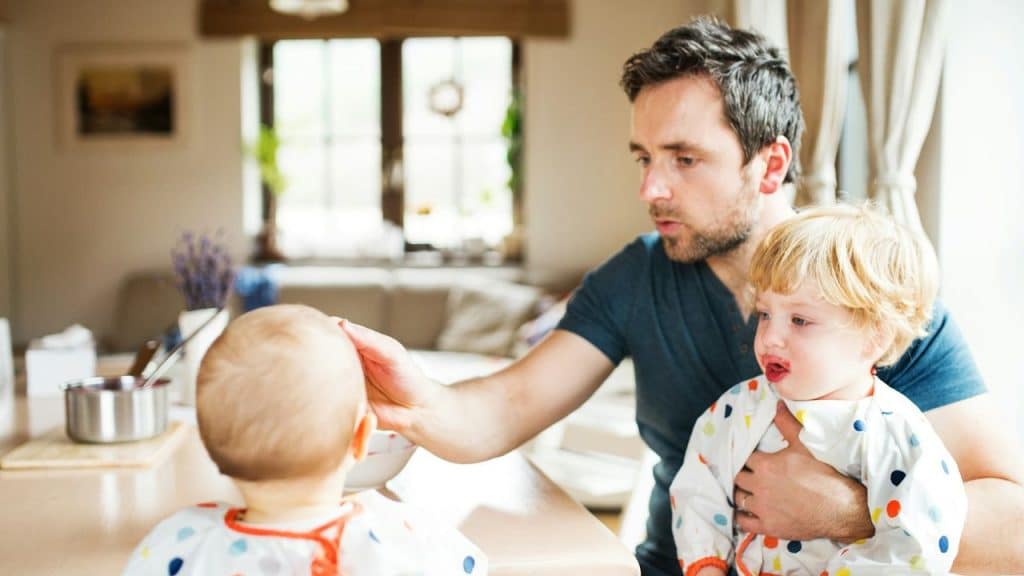 A man holds a baby in a bib while feeding a second baby in a bright, sunlit room.