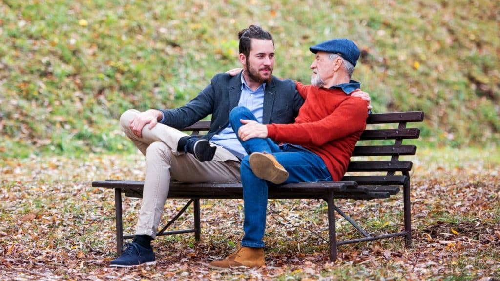 Two men of different ages sit on a park bench in autumn, with one's arm around the other.
