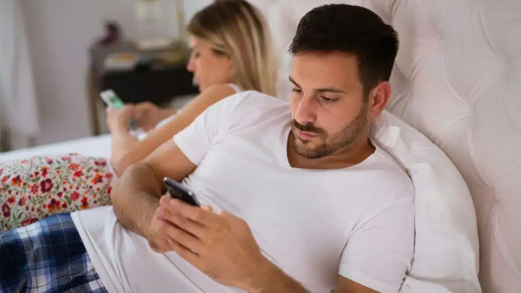 Man and woman lying in bed, both preoccupied with looking at their phones.