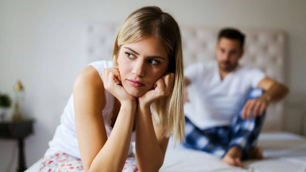 Blonde woman sitting in foreground, looking away; man in background sitting on a bed.