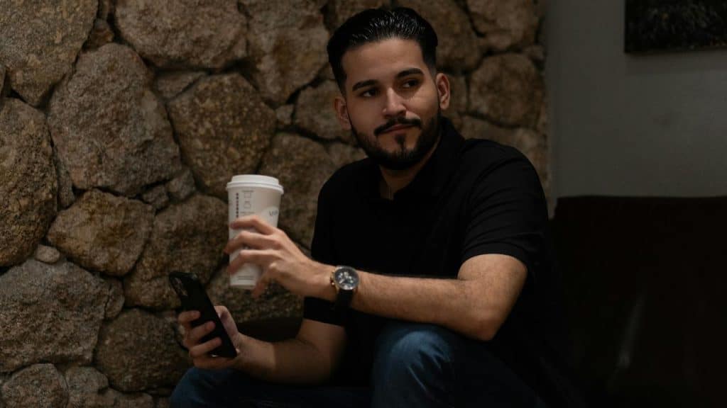 A man sitting indoors holding a coffee cup and a phone.
