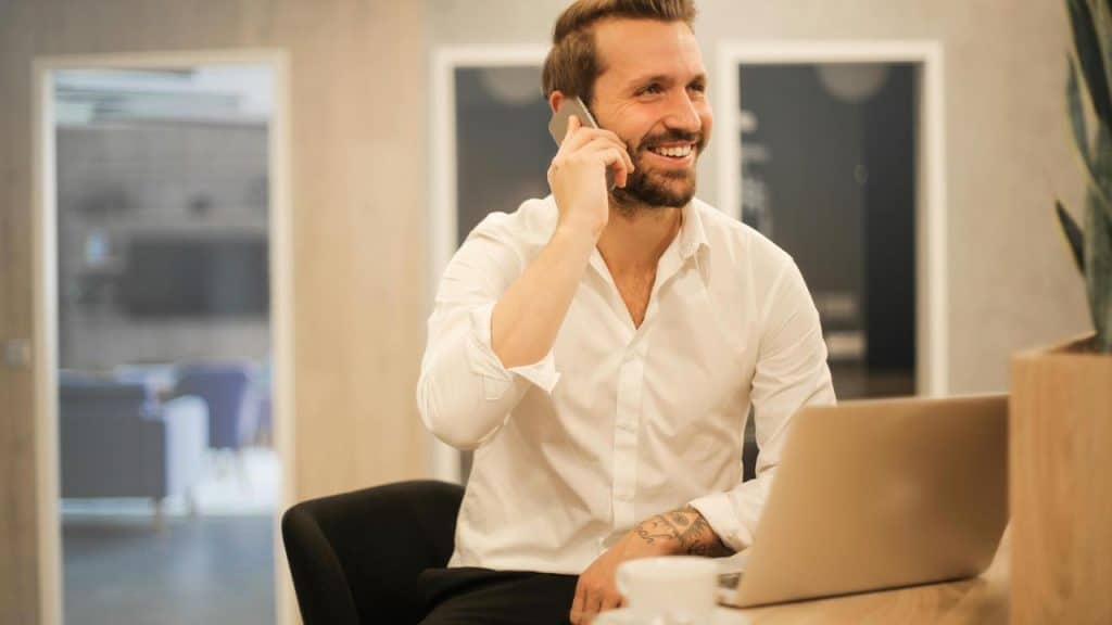 A man smiling while talking on the phone at a desk with a laptop.