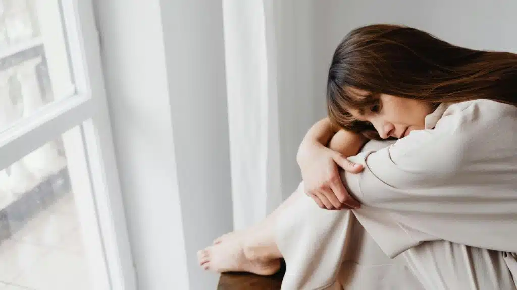 A woman sitting by a window, hugging her knees and appearing sad.
