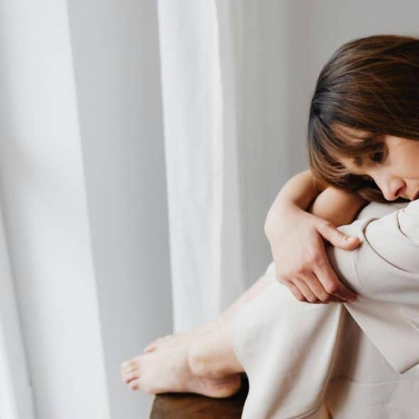 A woman sitting by a window, hugging her knees and appearing sad.
