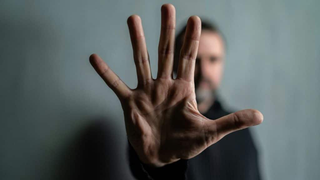 A blurry man holds his right hand out to the camera, palm forward, against a gray background.