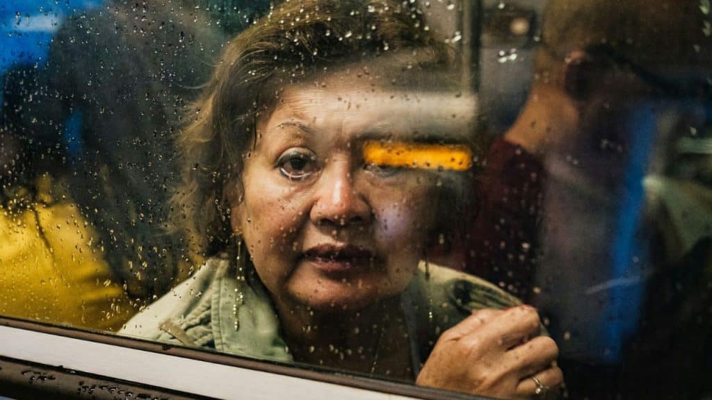 A woman looks through a window covered in rain drops, with an orange light streak across her eye.