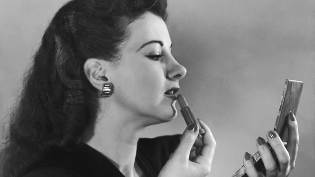 A black and white profile close-up of a woman applying lipstick while looking into a compact mirror.