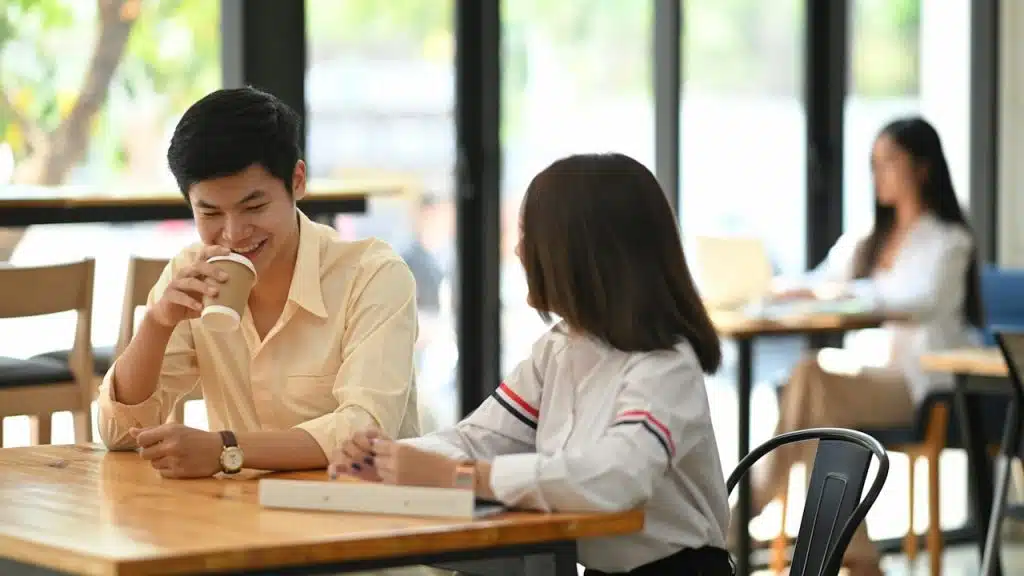A smiling young man drinking coffee and talking to a woman across a wooden table in a café.