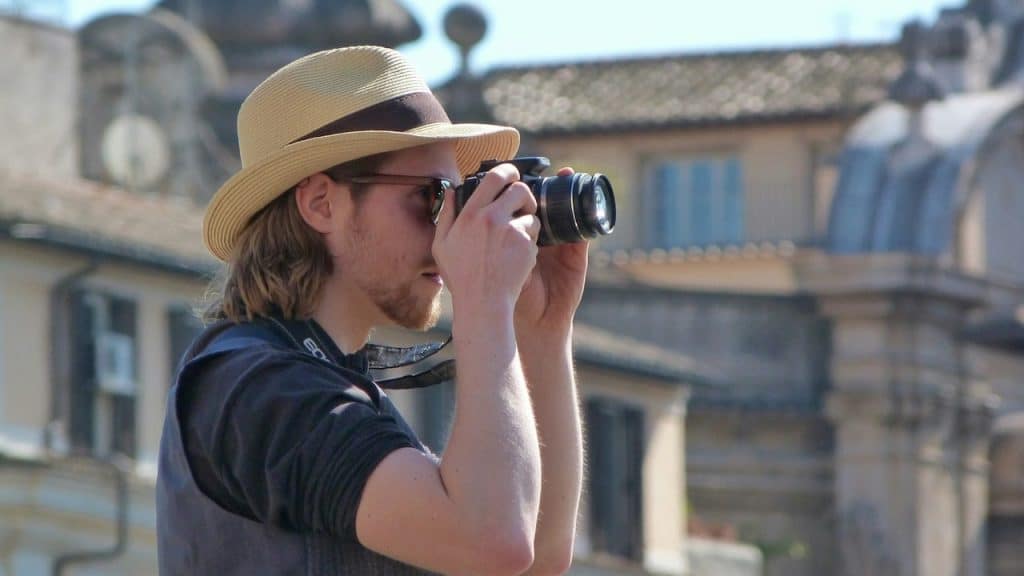 A man in a black t-shirt and brown fedora hat using black binoculars.