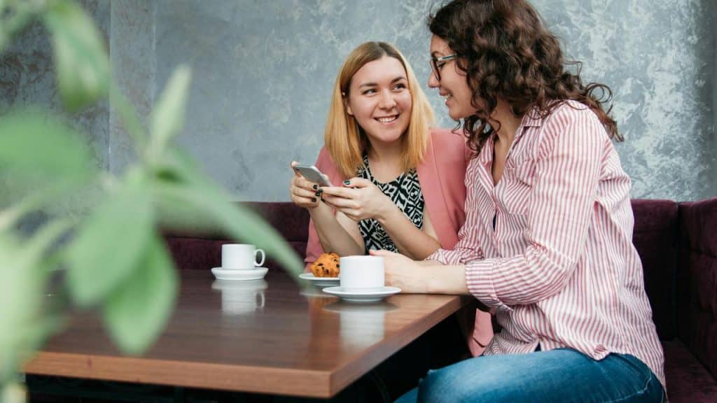 A pair of friends sitting at a café table smiling and talking over coffee.
