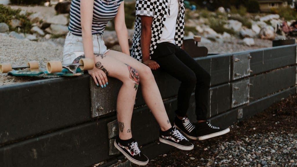 Two people, one with leg tattoos, sit on a low black wall next to skateboards.