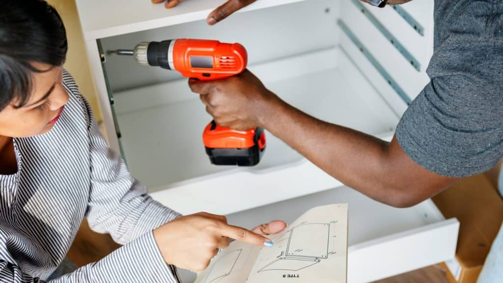 A man is installing a cabinet with an electric drill and his wife is helping him.