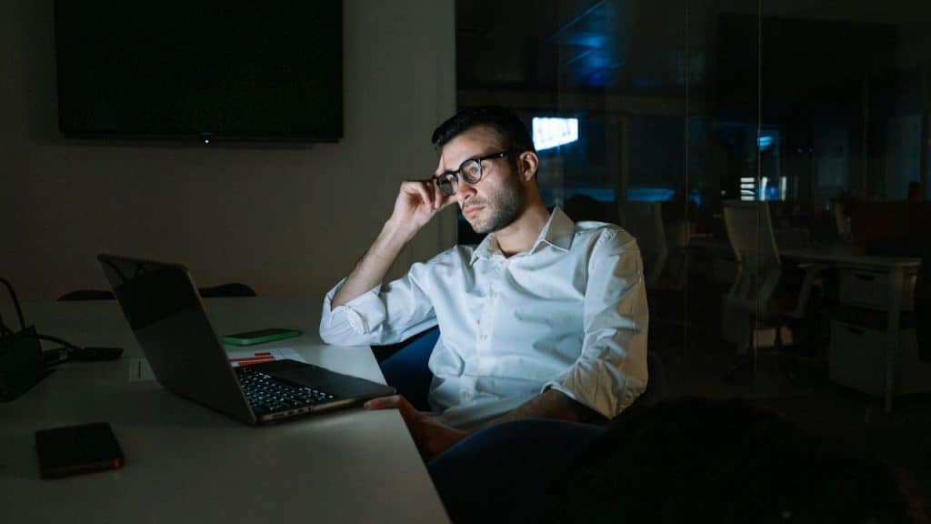 A man in a white shirt and glasses works late at a laptop in a dark office.