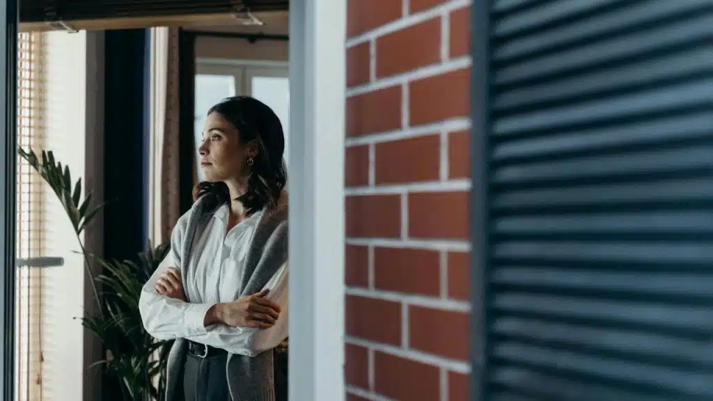 A person standing indoors looking out a window with arms crossed.