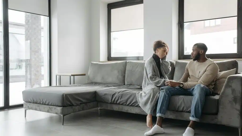 A couple sits on a couch holding hands and talking.