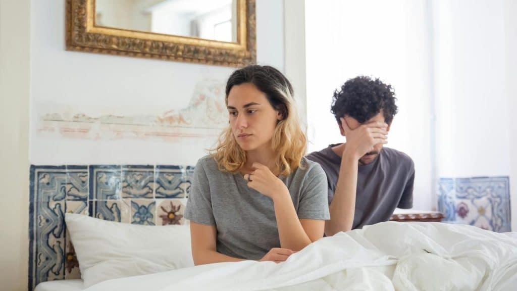 A couple sitting in bed, both looking upset and distant.