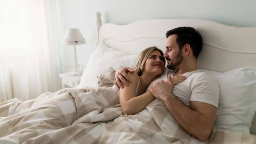 A man and woman are lying in bed, embracing and smiling at each other.