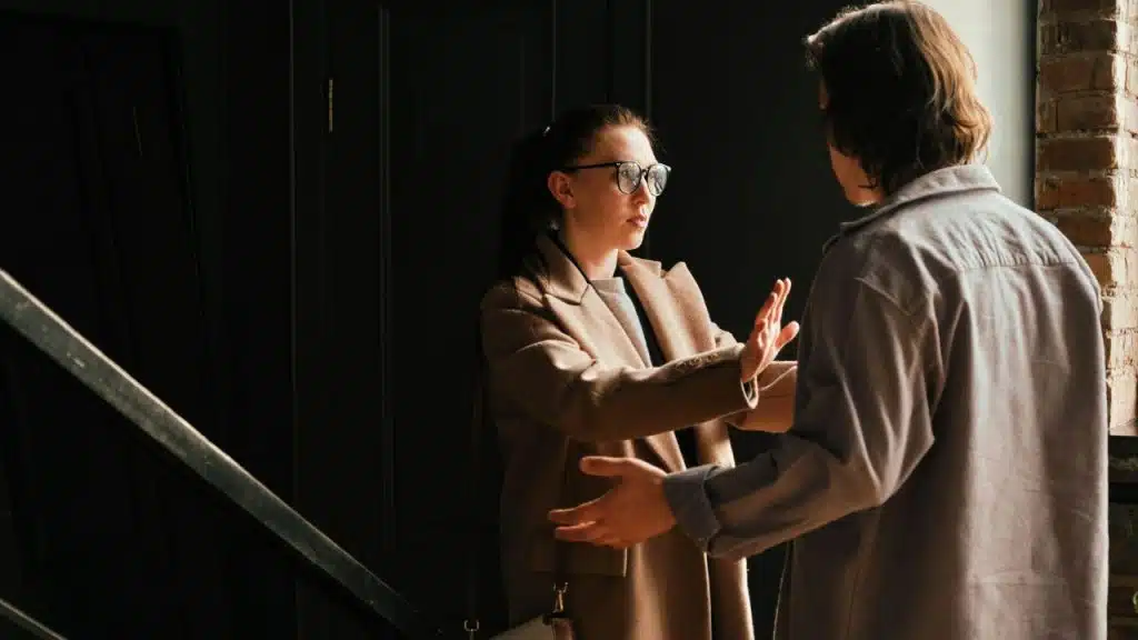 A woman in a tan coat holds up her hand to a man, suggesting he stop or step back.