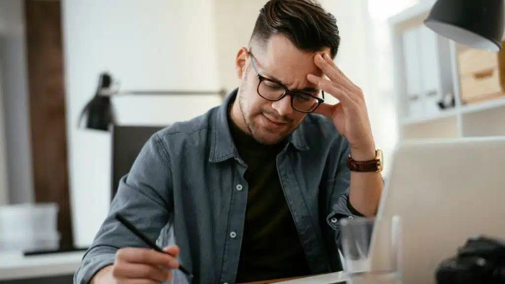 A distressed man wearing glasses is sitting at a desk, holding his head.