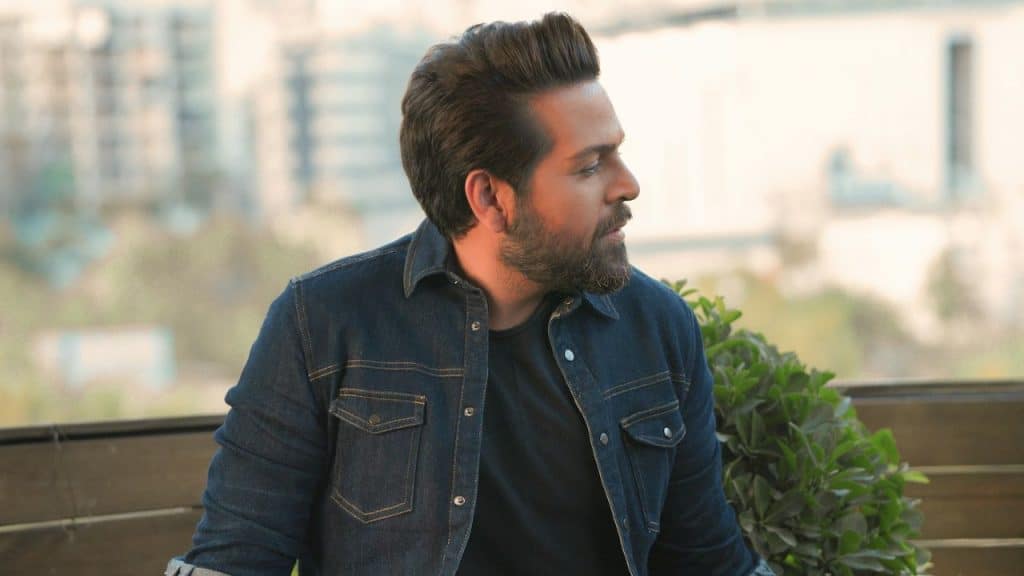 A bearded man in a denim jacket is looking to the side outdoors.