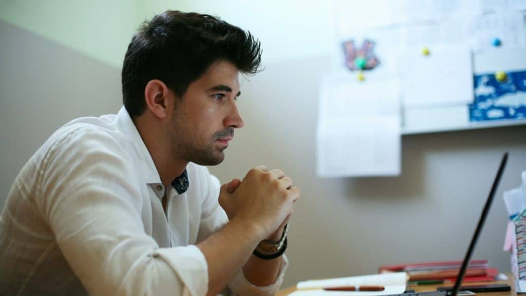 A focused man sitting at a desk with his hands clasped, looking at a laptop screen.