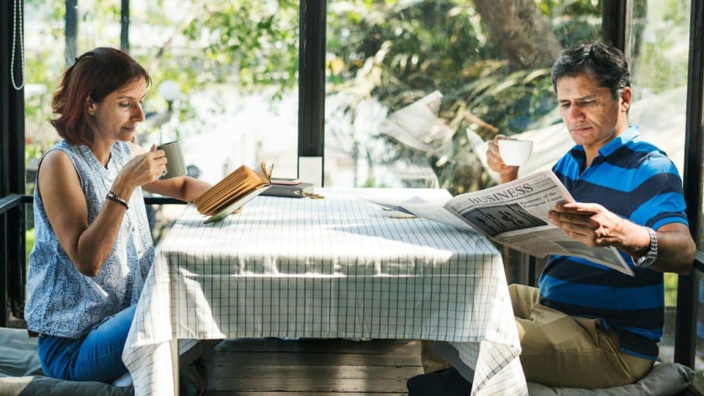 Man reading a newspaper and a woman reading a book, both holding coffee mugs.