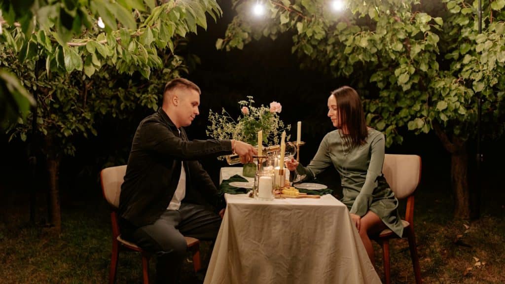 A couple sitting at a small table outdoors under string lights, clinking wine glasses during a romantic dinner.