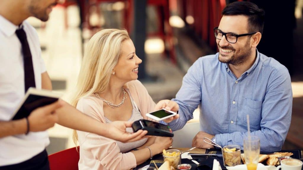 A smiling couple paying for their meal at an outdoor café.