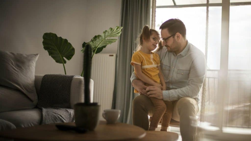A father holds a young girl in a yellow shirt while sitting near a window.