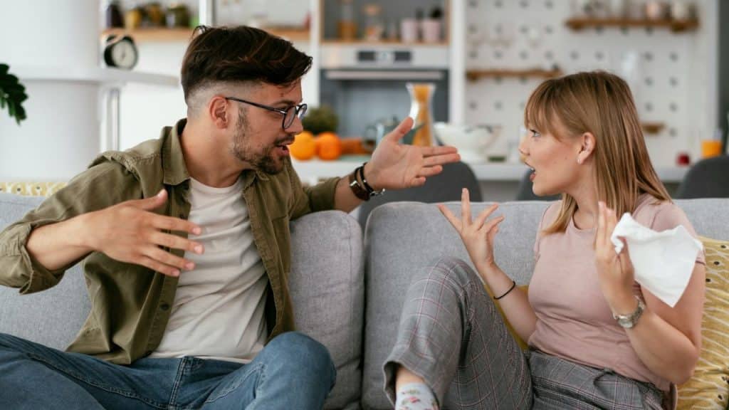 A man and woman sit on a couch facing each other, engaged in a heated argument with expressive hand gestures.