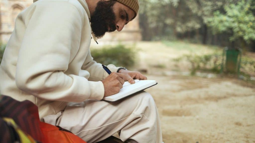 A bearded man in a beige hoodie and beanie writes in a notebook outdoors.