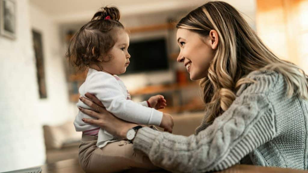 A smiling woman looks at a young child sitting on a table in a bright room.