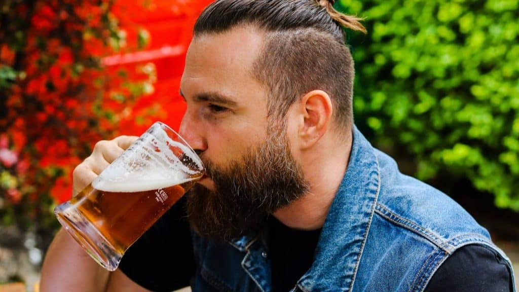 Man with a full beard and a man bun taking a drink from a glass mug of beer outdoors.