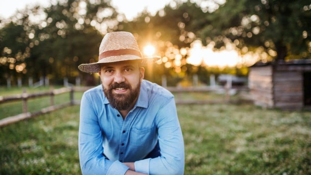 A bearded man in a straw hat smiles at the camera with the sunset behind him.