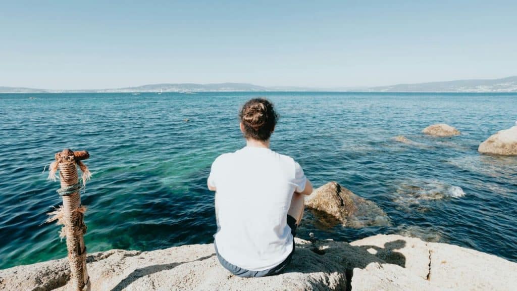 Person in a white shirt with a man bun sitting on a rock by the ocean, looking out at the water.