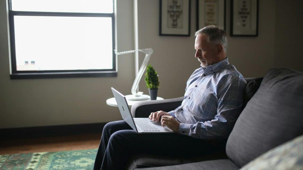 Middle-aged man in a plaid shirt sitting on a couch, using a laptop in a well-lit living room.